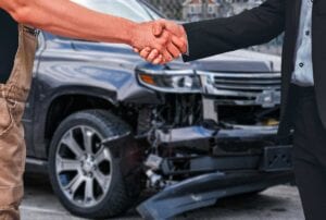 Mechanic And Professionally Dressed Customer Shake Hands In Front Of Damaged SUV